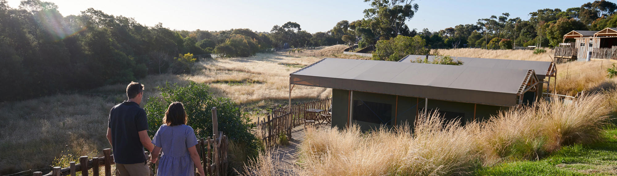 Two people with backs to camera walk hand-in-hand towards a hut at dusk.