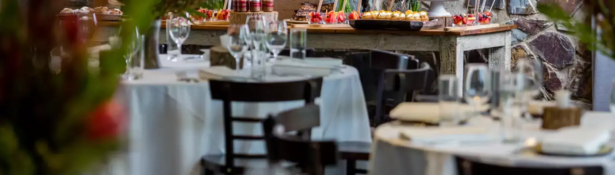 A number of tables with a white table cloth and glassware as well as a table in the background with sweet treats on it.