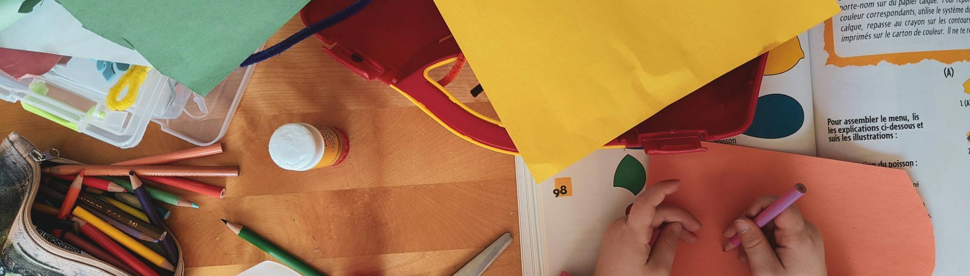 A top-down view of a child's desk featuring assorted coloured paper, pencils, glue, a bottle, scissors, felt wire, a sharpener and a (French) papercraft activity book.
