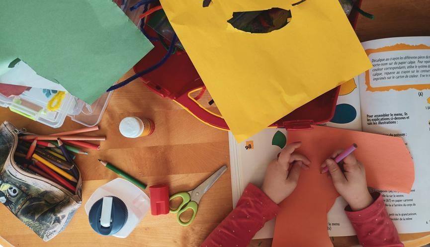 A top-down view of a child's desk featuring assorted coloured paper, pencils, glue, a bottle, scissors, felt wire, a sharpener and a (French) papercraft activity book.