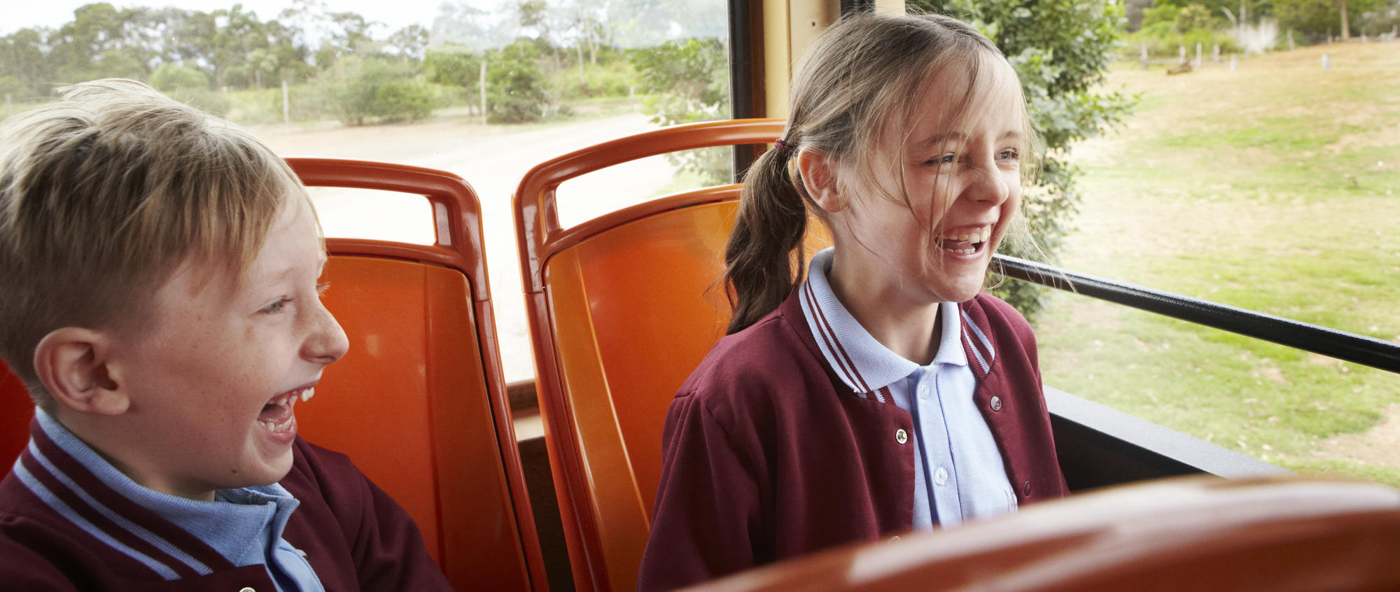 Two young guests sitting in the back of a Safari Bus, both looking right and laughing joyfully.
