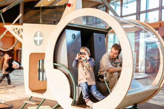 A boy is in a play helicopter inside looking through binoculars. A man in a zoo keeper uniform is sitting next to him.