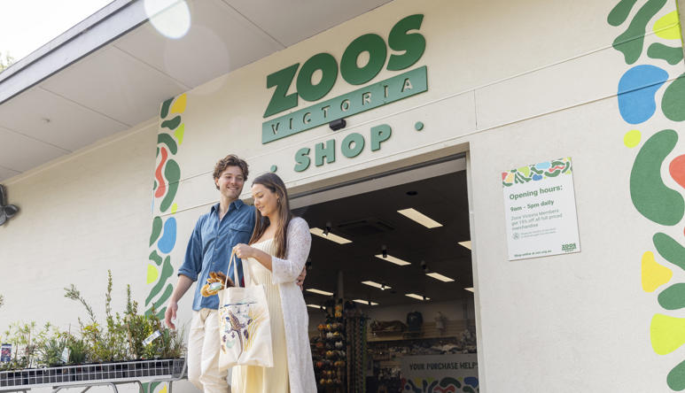 Two shoppers exit the Melbourne Zoo Shop, facing right, with a plush Giraffe in the tote bag.