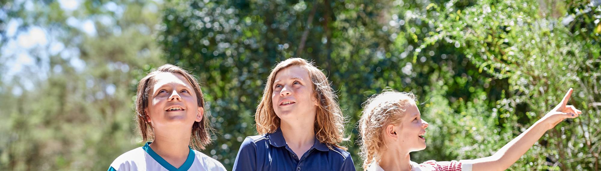 Three students smile as they walk through the Sanctuary bush on a sunny day, looking and pointing in various directions.