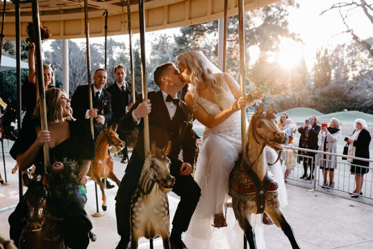 Wedding ceremony on the Carousel, where at least nine guests bear witness to the newlyweds kissing while sitting on horses.