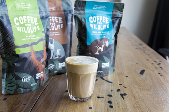 Three "Coffee For Wildlife" bags behind a full latte glass on a wooden table.