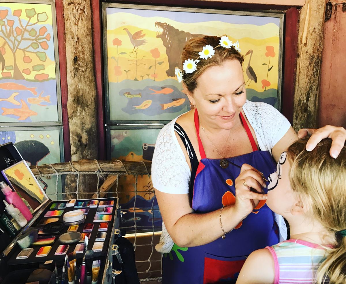 A woman wearing a flower crown is leaning over a child and painting their face with face paint.