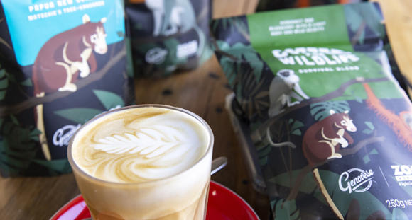 A milky coffee in a glass with latte art in the froth; the coffee sits on a saucer with a teaspoon, in front of bags of coffee on a table.