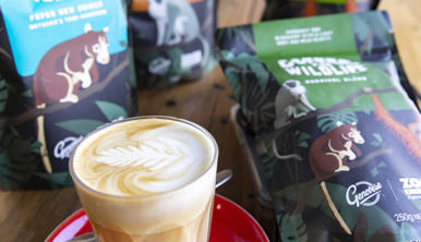 A milky coffee in a glass with latte art in the froth, sitting on a saucer with a teaspoon, in front of bags of coffee on a table.