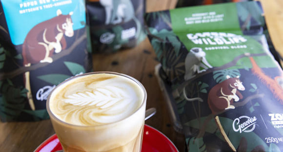 A milky coffee in a glass with latte art in the froth, sitting on a saucer with a teaspoon, in front of bags of coffee on a table.