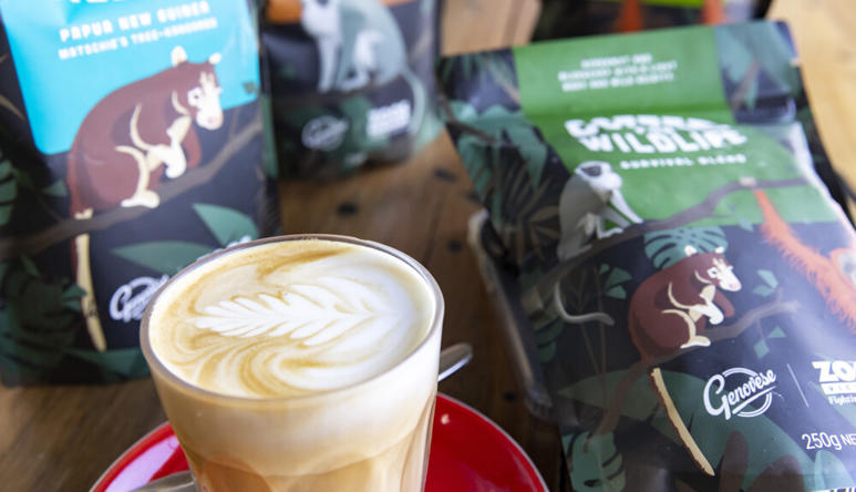A milky coffee in a glass with latte art in the froth, sitting on a saucer with a teaspoon, in front of bags of coffee on a table.