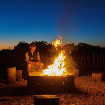 A camp fire billows at dusk. A man is smiling, sitting behind the campfire.