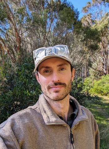 Portrait of Biologist Darcy, in front of a wooded backdrop, wearing a cap holding a pair of clear safety goggles.