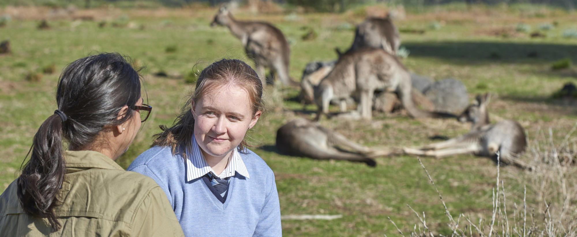 A student in light blue school uniform has her back to a pack of Kangaroos in a field as she talks with a Keeper in khaki uniform.