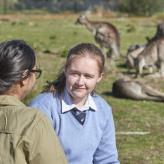 A student in light blue school uniform has her back to a pack of Kangaroos in a field as she talks with a Keeper in khaki uniform.