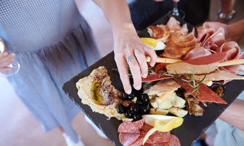 Overhead shot of a platter containing biscuits, salami, cheese and other antipasto; A hand is reaching for an olive on the platter.