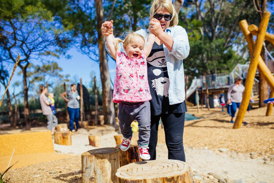 A young girl is stepping across some logs while a woman holds her hands up and helps her balance.