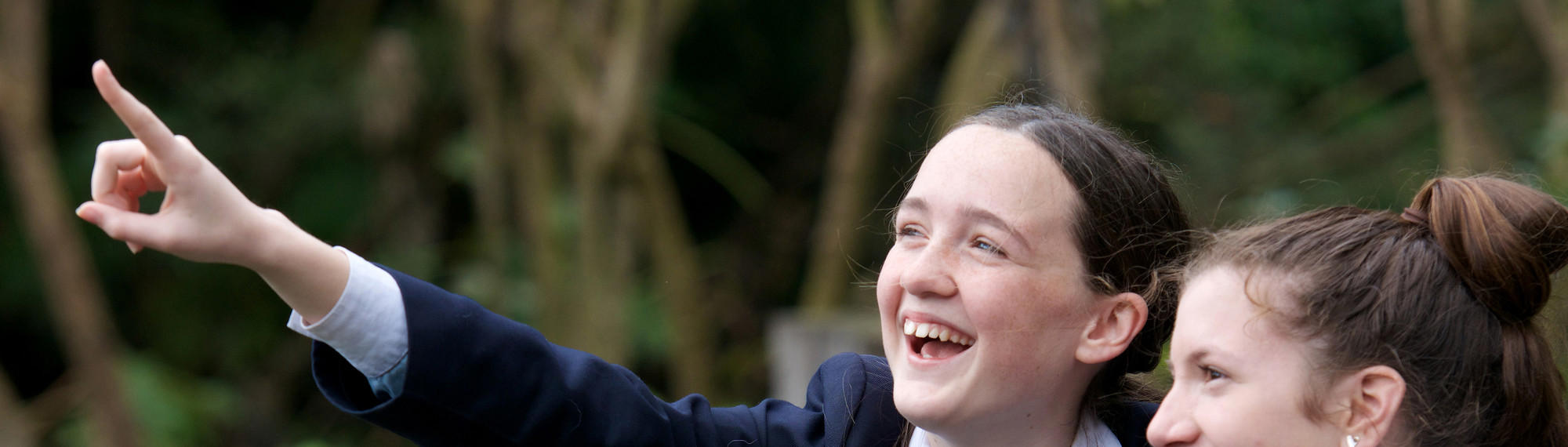 Two teen girls dressed in school uniform look excitedly at something out of frame, one pointing left.