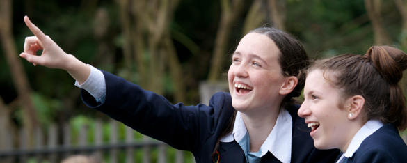 Two teen girls dressed in school uniform look excitedly at something out of frame, one pointing left.