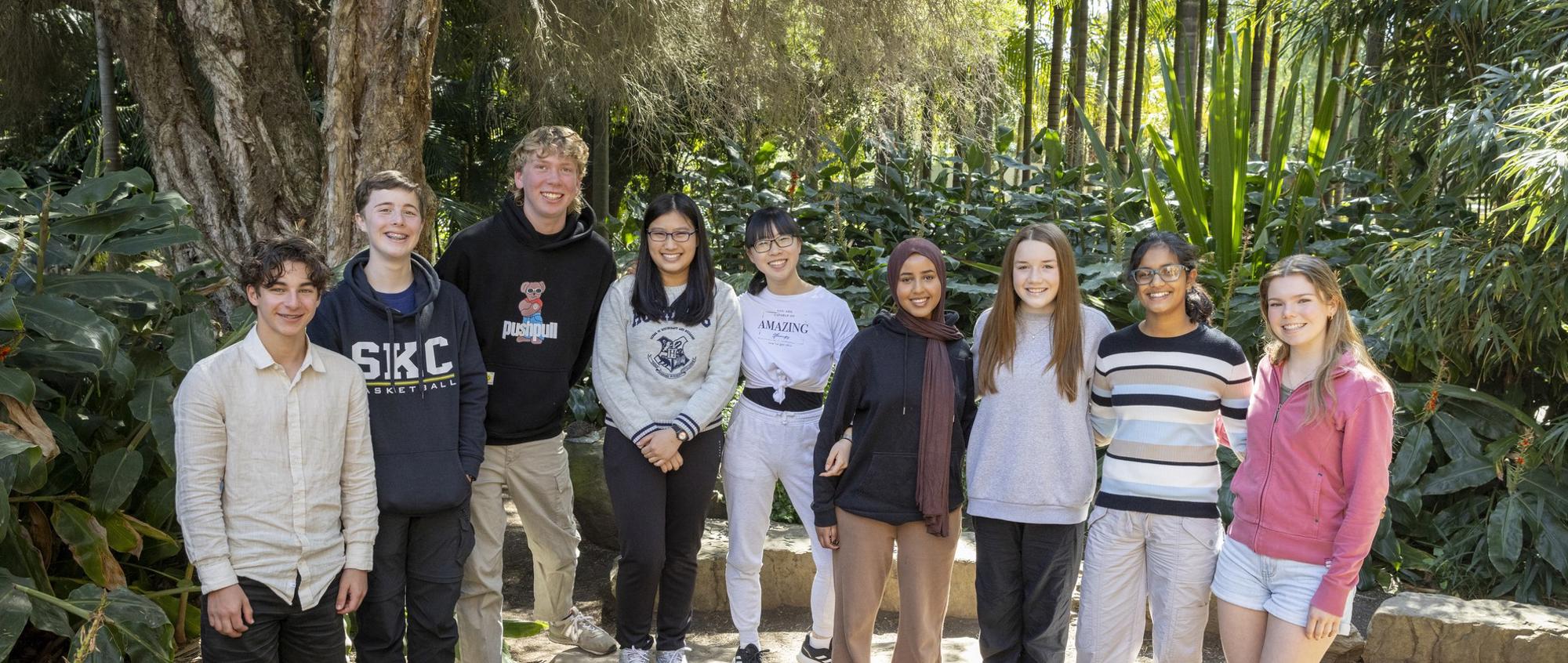 A group of nine teenage students standing in a ground and smiling, in front of a rainforest setting.