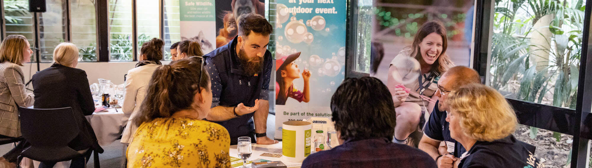 A group of four visitors sit around a table learning from a Zoos Victoria staff member, with "Blow Bubbles" posters beside them.