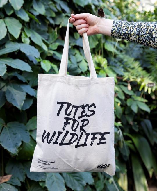 A tote bag with the text "Totes for Wildlife" on it is held up in front of a wall of tropical plants; a butterfly has landed on the hand holding the bag.