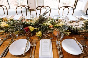 View of wedding banquet table, set up with crockery, cutlery, glasses, flowers and a custom menu.