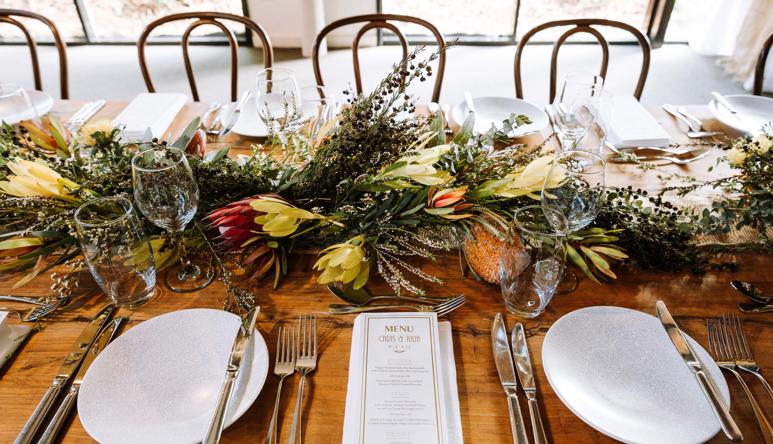 View of wedding banquet table, set up with crockery, cutlery, glasses, flowers and a custom menu.