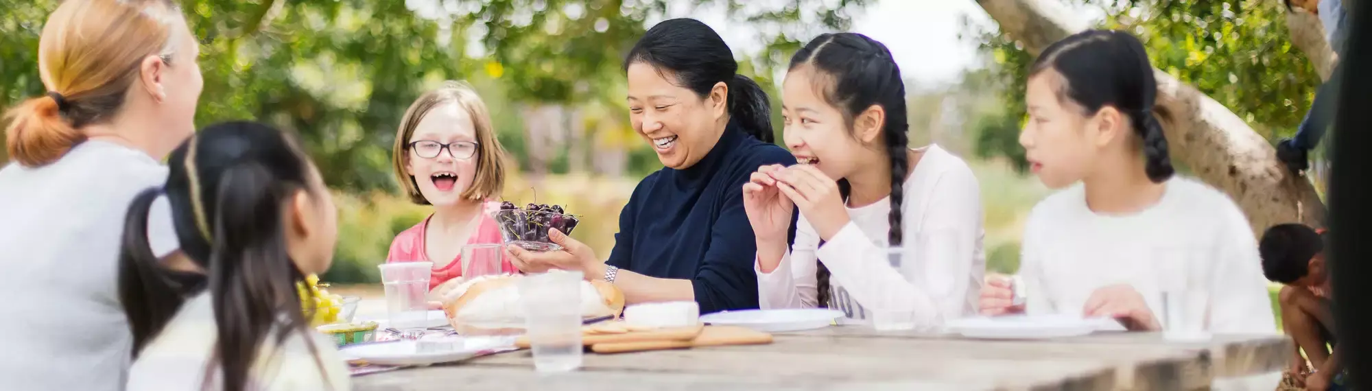 A group of adults and children are sitting at an outdoor table laughing; One woman is holding a bowl of cherries.