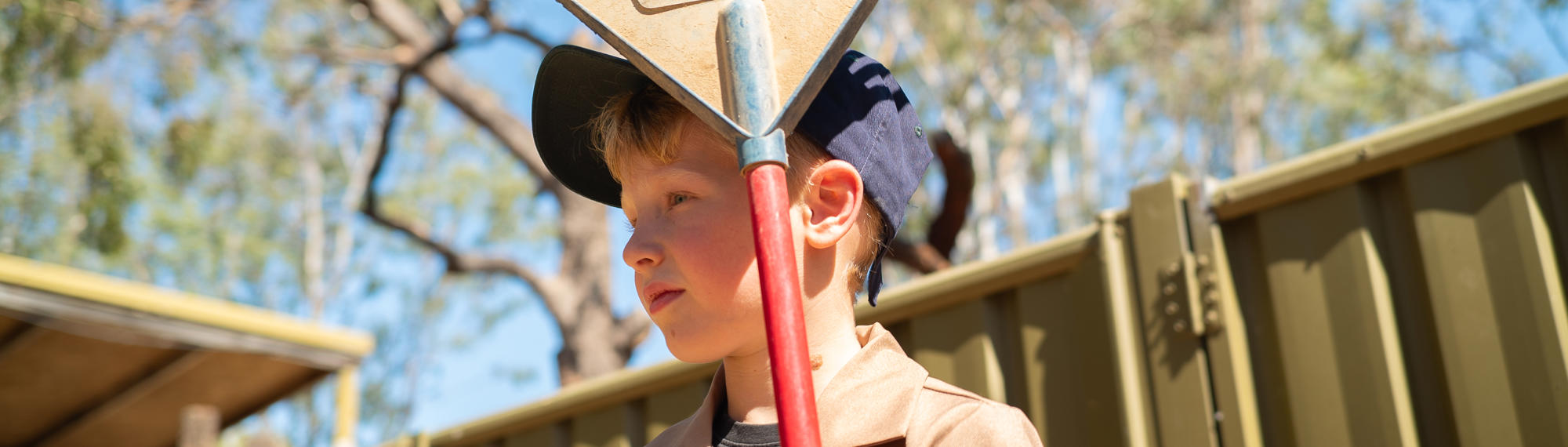 Junior "Zoo-Keeper" holding a rake, with red handle and dust-covered blue head, looking left and standing in front of a corrugated olive yellow fence.