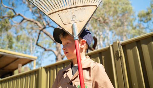 Junior "Zoo-Keeper" holding a rake, with red handle and dust-covered blue head, looking left and standing in front of a corrugated olive yellow fence.