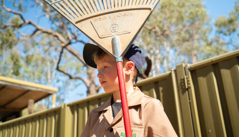 Junior "Zoo-Keeper" holding a rake, with red handle and dust-covered blue head, looking left and standing in front of a corrugated olive yellow fence.