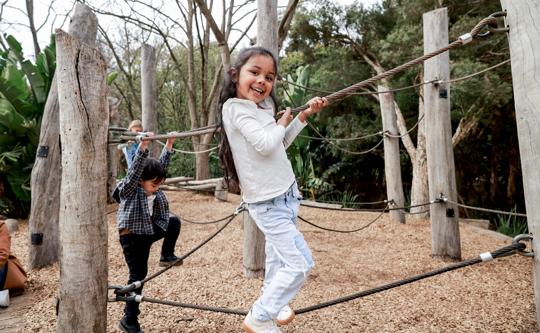 A young girl is smiling as she balances on a rope and holds onto another rope in a playground.